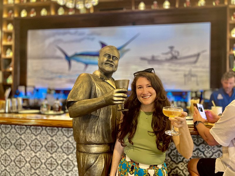 Guest raising a cocktail beside the bronze Ernest Hemingway statue inside the Hemingway Distillery's Social Club in Key West