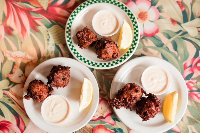Three plates of golden conch fritters with dipping sauce and lemon wedges on a tropical floral tablecloth
