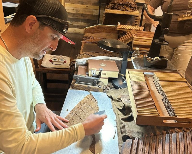 Danny Rodriguez trimming a tobacco leaf at Rodriguez Cigar Factory, his workbench lined with finished hand-rolled cigars