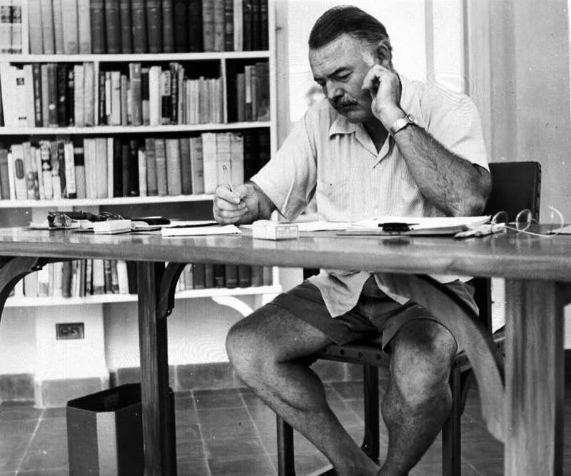 Archival photograph of Ernest Hemingway writing at his desk in front of a wall of books