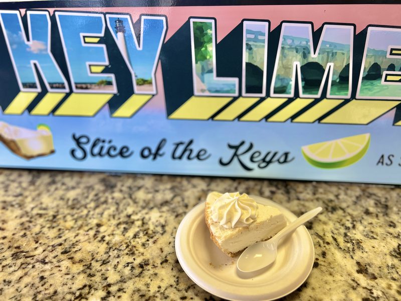 Slice of Key lime pie topped with whipped cream in front of a Key Lime Pie Company sign in Key West