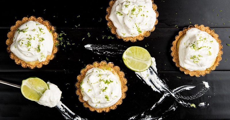 Four mini Key lime tarts topped with whipped cream and lime zest on a dark surface