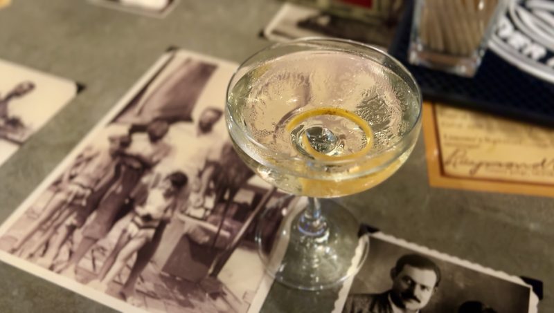 A coupe cocktail with a citrus twist resting on a table spread with vintage black-and-white Hemingway-era photographs at the Pilar Social Club