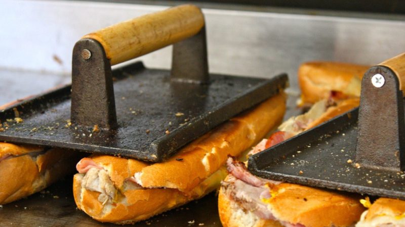 Pressed Cuban sandwiches from Cuban Coffee Queen under cast-iron plancha weights on a hot griddle