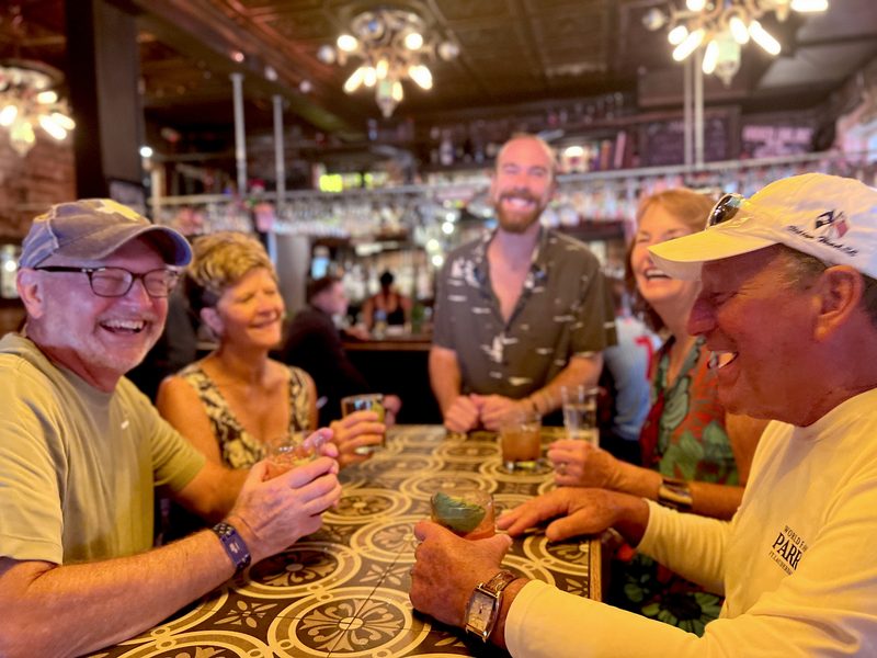 Tour group laughing over cocktails at General Horseplay in Key West, under a pressed-tin ceiling and chandeliers