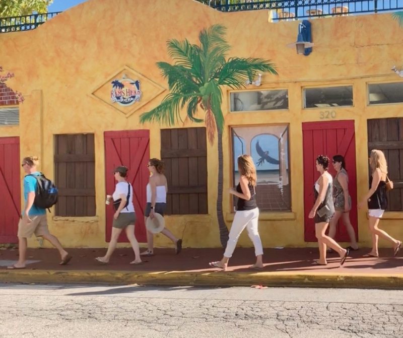 Tour group walking single file along Petronia Street in Bahama Village, past a yellow building painted with a palm mural