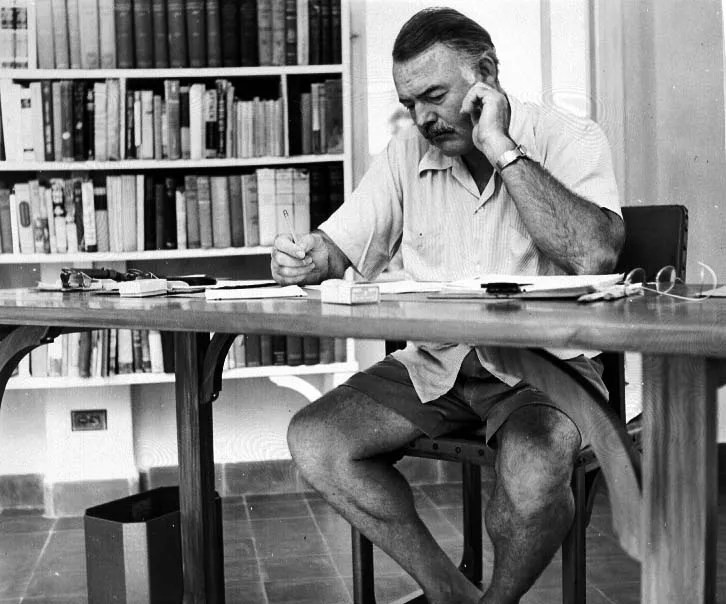 Ernest Hemingway sitting on a bench in front of a book shelf